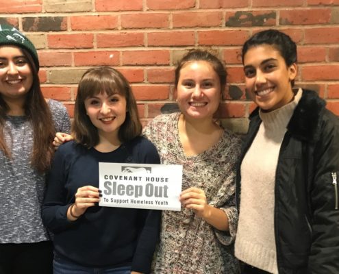 Four teenage girls stand smiling in front of a brick wall. The middle two girls hold each side of a piece of paper reading "Covenant House Sleep Out to Support Homeless Youth".