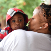 A mother with her back to the camera holds her baby while looking at him. The infant is looking over her shoulder into the camera with wide eyes.