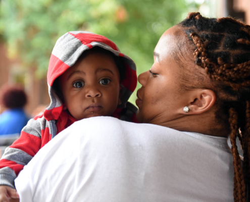 A mother with her back to the camera holds her baby while looking at him. The infant is looking over her shoulder into the camera with wide eyes.