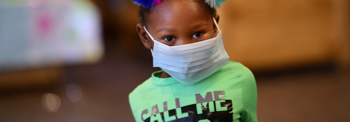 A child wears a mask that is too large for their face while looking at the camera