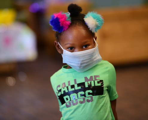 A child wears a mask that is too large for their face while looking at the camera