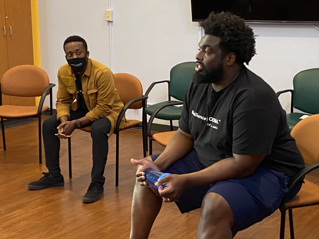 Two men sit in chairs. One is wearing a mask, looking at the other one who is speaking. The man speaking has a shirt that reads, 