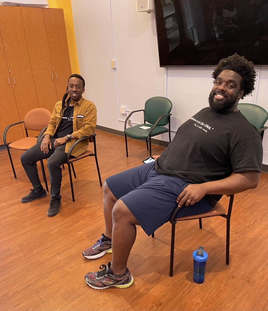 Two men sit in chairs, smiling at the camera. The chairs are on hard wood floor.