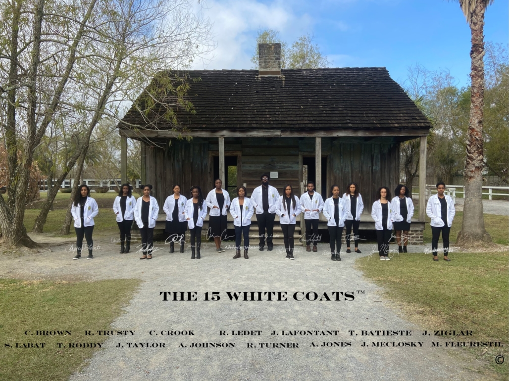 Fifteen people in white coats stand in front of a wooden house in a line