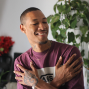 A young man in a maroon t-shirt is smiling with his eyes closed, crossing his arms over his chest. He stands in front of a plant.