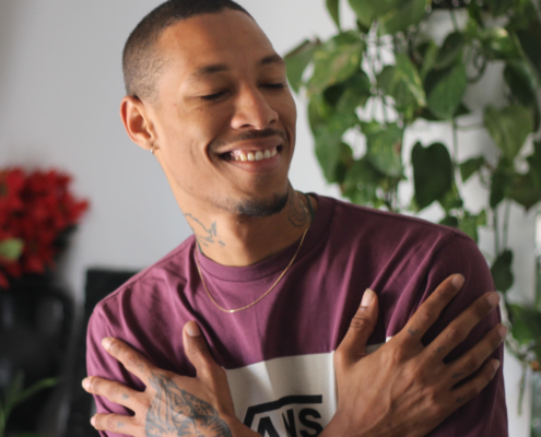 A young man in a maroon t-shirt is smiling with his eyes closed, crossing his arms over his chest. He stands in front of a plant.